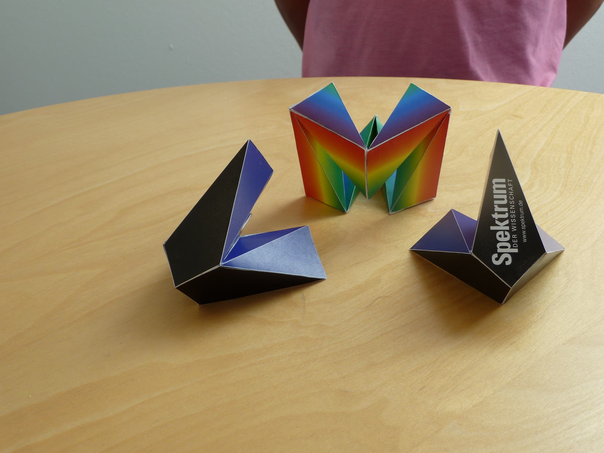 Drei bunte, geometrische Papiermodelle stehen auf einem Holztisch. Das mittlere Modell zeigt ein Regenbogenmuster, während das rechte Modell den Text "Spektrum der Wissenschaft" trägt. Im Hintergrund ist eine Person in einem rosa T-Shirt zu sehen, deren Oberkörper teilweise sichtbar ist.