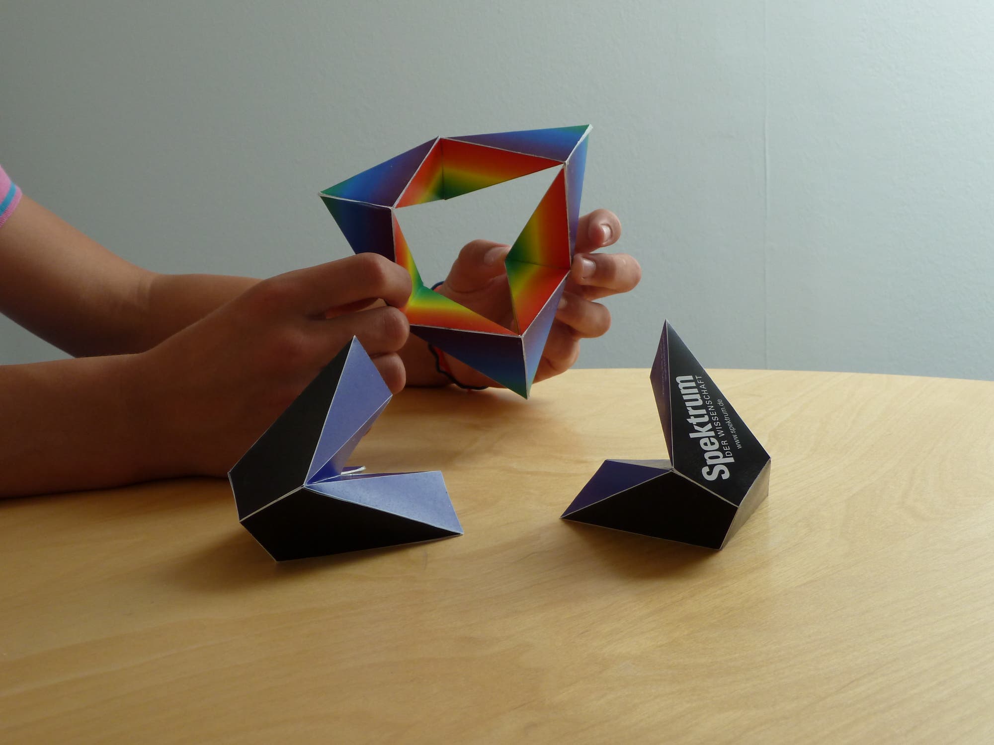 Eine Person hält ein buntes, geometrisches Papierobjekt in den Händen. Auf dem Tisch liegen zwei weitere ähnliche Objekte. Eines der Objekte zeigt den Text "Spektrum". Die Szene spielt sich auf einem hellen Holztisch vor einer neutralen Wand ab.