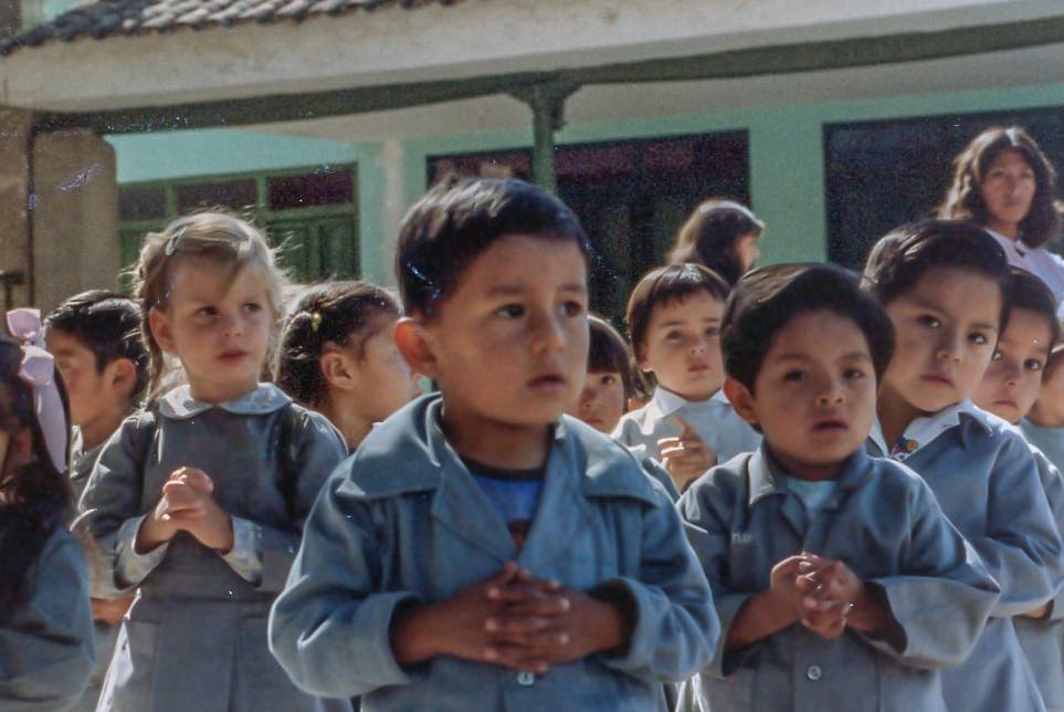 Eine Gruppe von Kindern in Schuluniformen steht in einer Reihe im Freien. Sie haben die Hände gefaltet und blicken aufmerksam nach vorne. Im Hintergrund ist ein Gebäude mit grünen Wänden zu sehen. Eine Frau steht im Hintergrund und beobachtet die Kinder. Die Szene wirkt wie eine Schulveranstaltung oder ein Morgenappell.