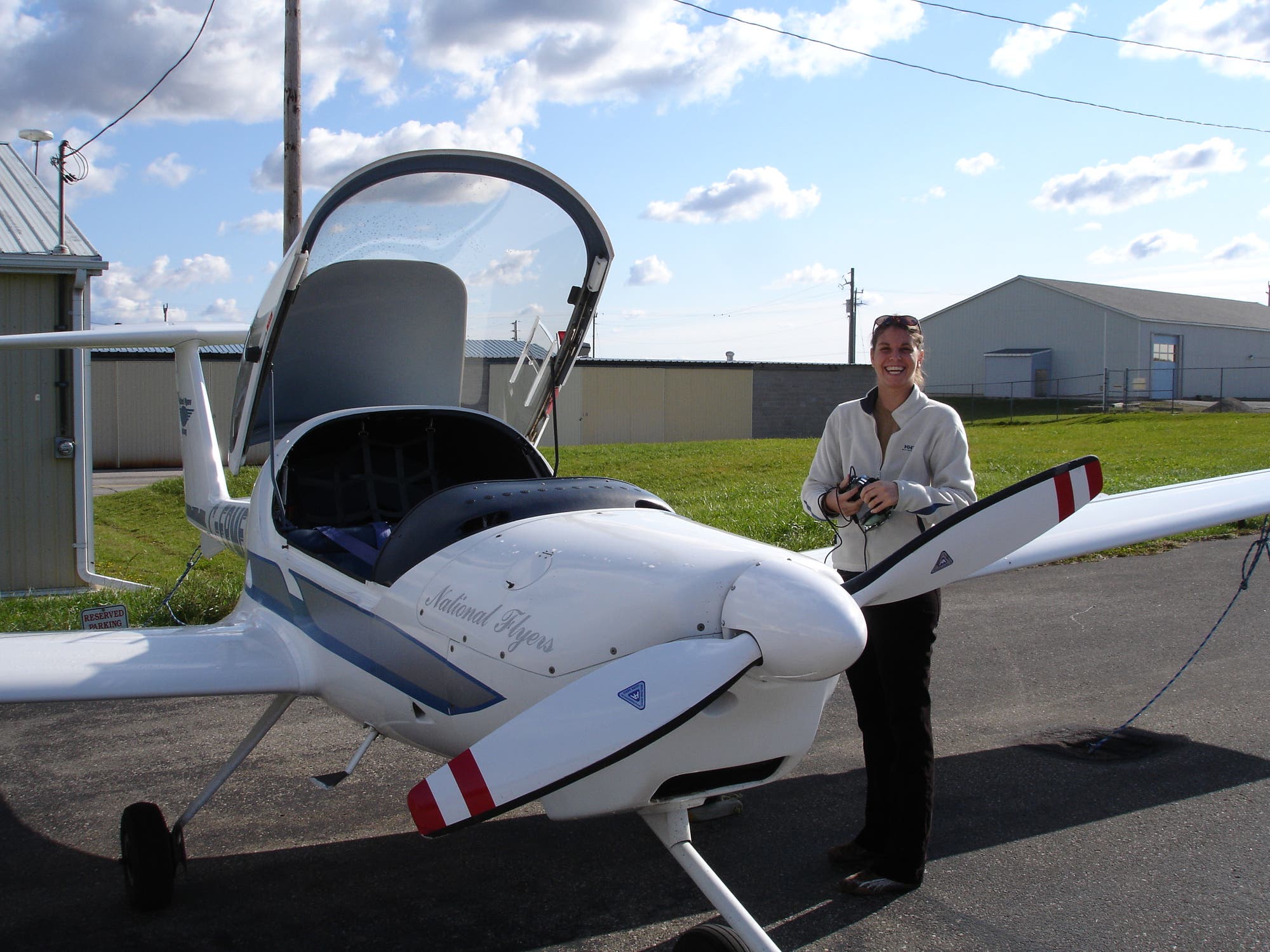 Eine Person steht lächelnd neben einem kleinen weißen Flugzeug mit geöffneter Cockpithaube auf einem Flugplatz. Auf dem Flugzeug steht "National Flyers". Im Hintergrund sind ein Hangar und ein grüner Rasen zu sehen. Der Himmel ist leicht bewölkt.