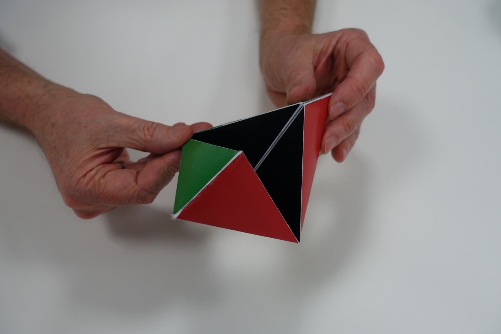 Zwei Hände halten ein dreidimensionales geometrisches Modell mit dreieckigen Flächen in den Farben Rot, Schwarz und Grün. Das Modell hat eine pyramidenartige Form. Der Hintergrund ist weiß.