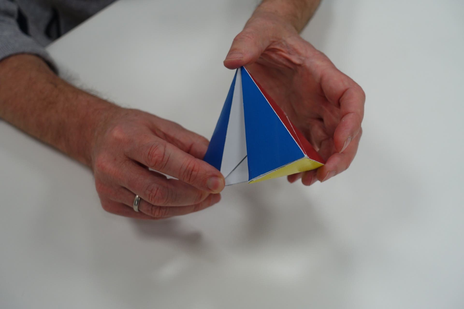 Zwei Hände halten ein buntes, dreieckiges Origami-Modell. Das Modell hat blaue, rote und gelbe Flächen. Die Person trägt einen silbernen Ring am linken Ringfinger. Der Hintergrund ist ein weißer Tisch.