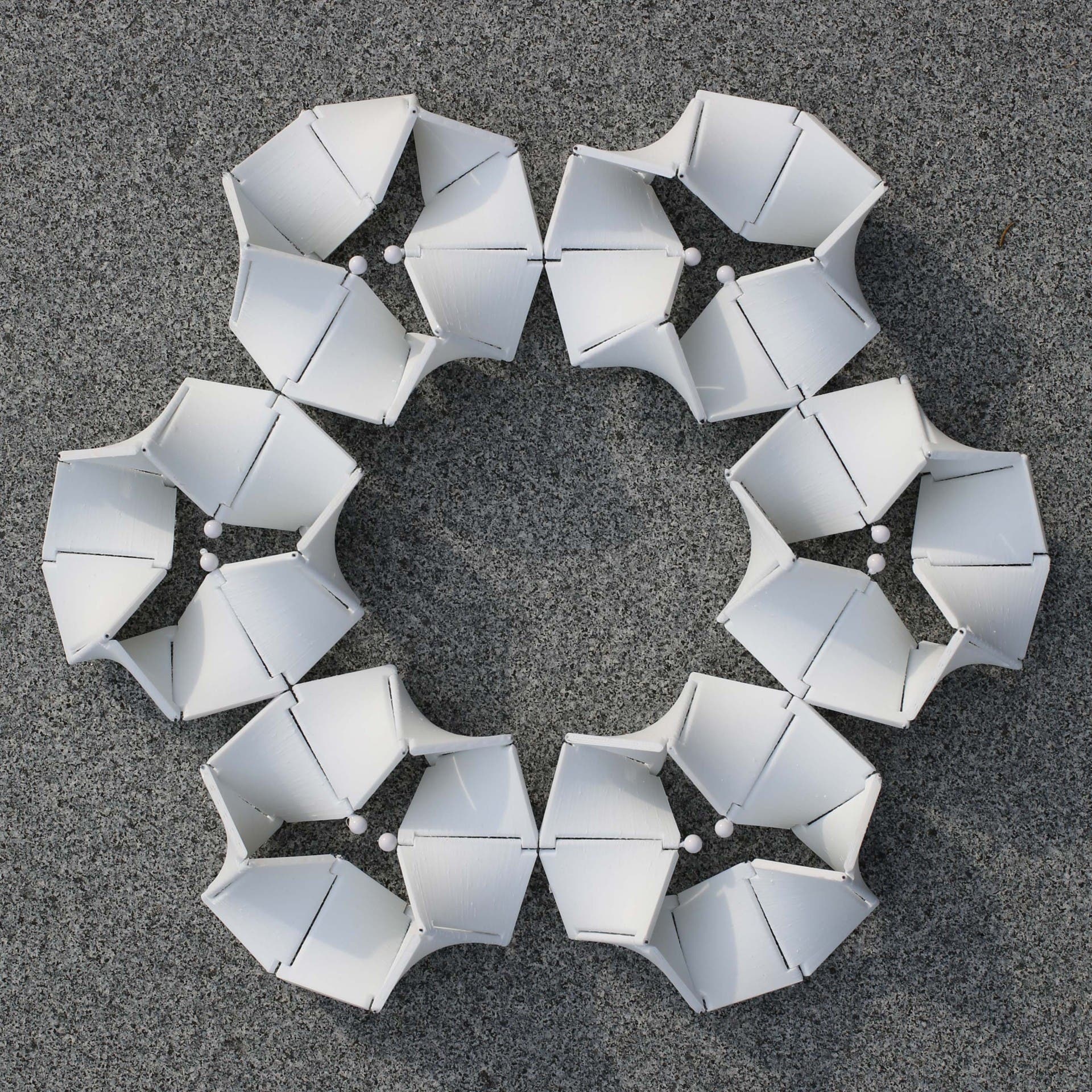 Eine kreisförmige Anordnung aus weißen, geometrischen Stühlen auf grauem Hintergrund. Die Stühle sind in einem symmetrischen Muster angeordnet, das an ein Schneeflocken-Design erinnert. Keine Personen sind im Bild zu sehen.