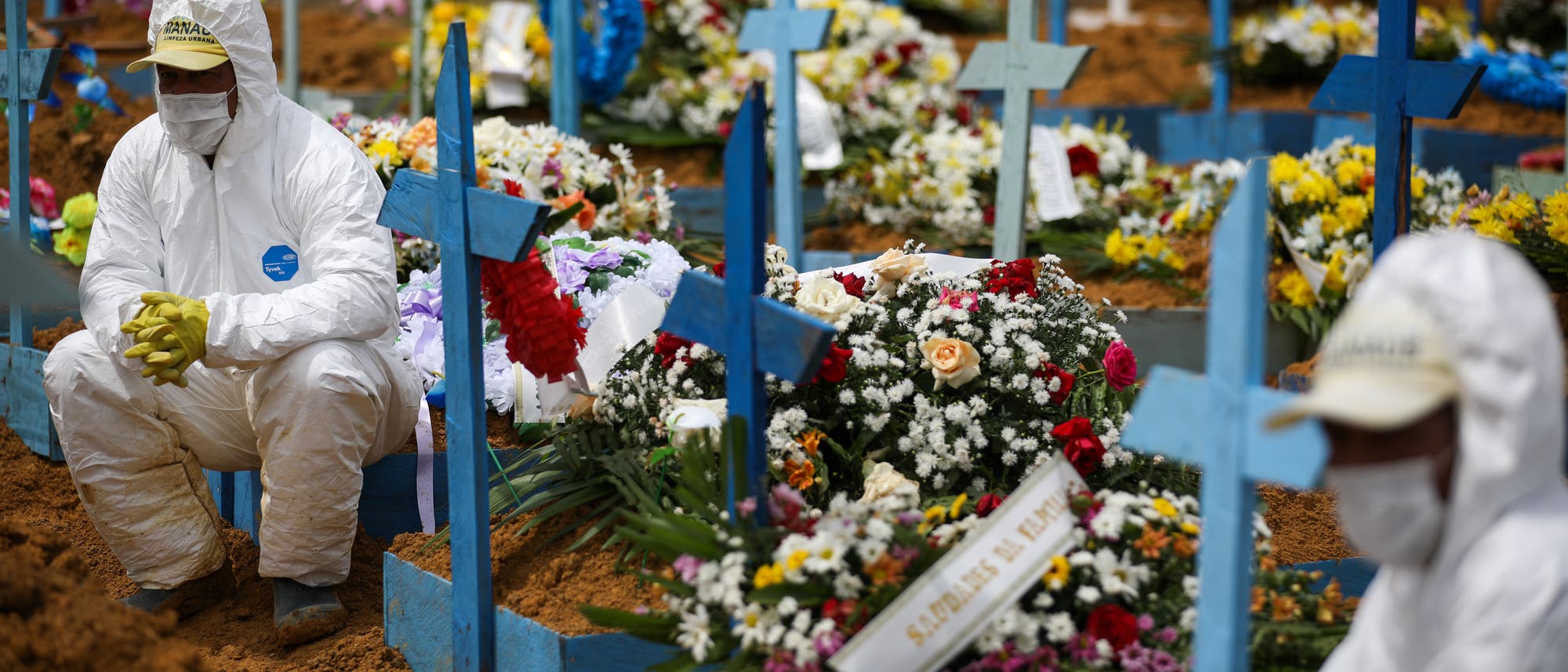 Friedhofsmitarbeiter in Schutzanzügen sitzen auf mit Blumen geschmückten Gräbern während einer Beerdigung am Friedhof Nossa Senhora Aparecida in Manaus. Friedhofsmitarbeiter in Schutzanzügen sitzen auf mit Blumen geschmückten Gräbern während einer Beerdigung am Friedhof Nossa Senhora Aparecida in Manaus.