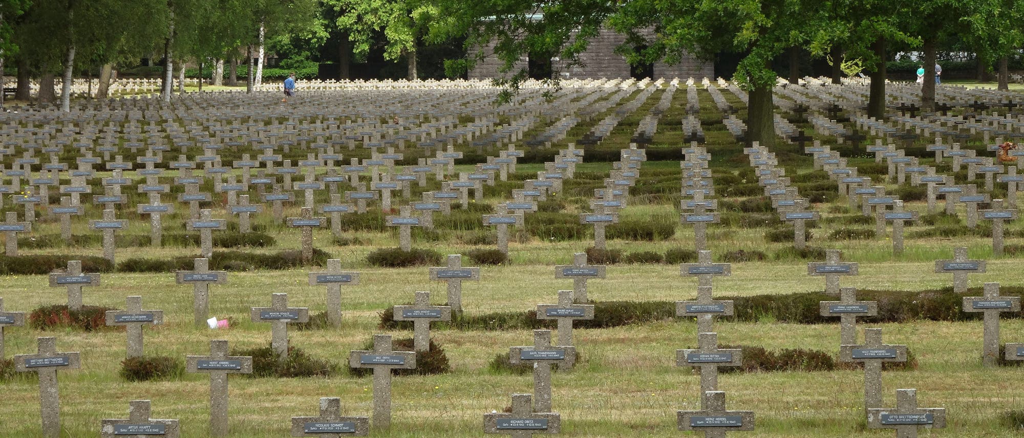 Ein weitläufiger Soldatenfriedhof mit zahlreichen grauen Steinkreuzen, die in geordneten Reihen aufgestellt sind. Im Hintergrund sind grüne Bäume zu sehen. Eine einzelne Person in blauer Kleidung geht zwischen den Gräbern entlang. Die Szene vermittelt eine ruhige und nachdenkliche Atmosphäre.