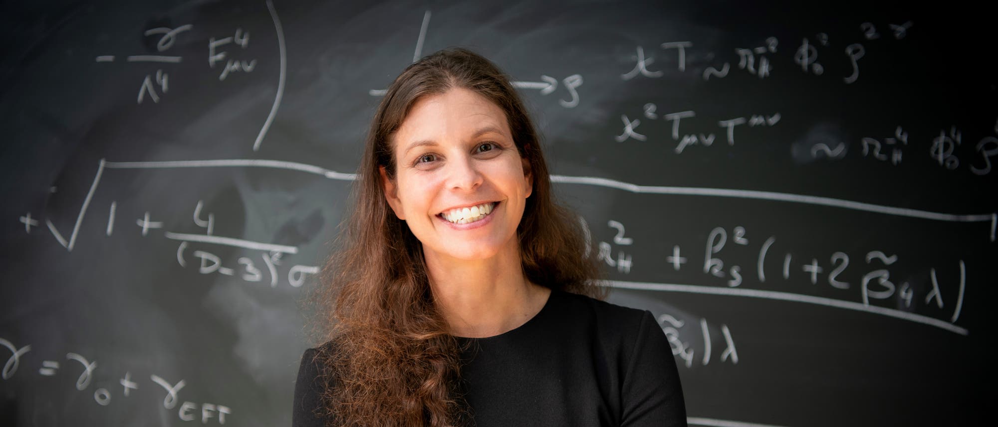 Eine lächelnde Frau steht vor einer Tafel, die mit komplexen mathematischen Formeln beschrieben ist. Sie trägt ein schwarzes Oberteil und hält ein Stück Kreide in der Hand. Die Tafel zeigt Gleichungen und Symbole, die auf fortgeschrittene Mathematik oder Physik hindeuten. Eine lächelnde Frau steht vor einer Tafel, die mit komplexen mathematischen Formeln beschrieben ist. Sie trägt ein schwarzes Oberteil und hält ein Stück Kreide in der Hand. Die Tafel zeigt Gleichungen und Symbole, die auf fortgeschrittene Mathematik oder Physik hindeuten.