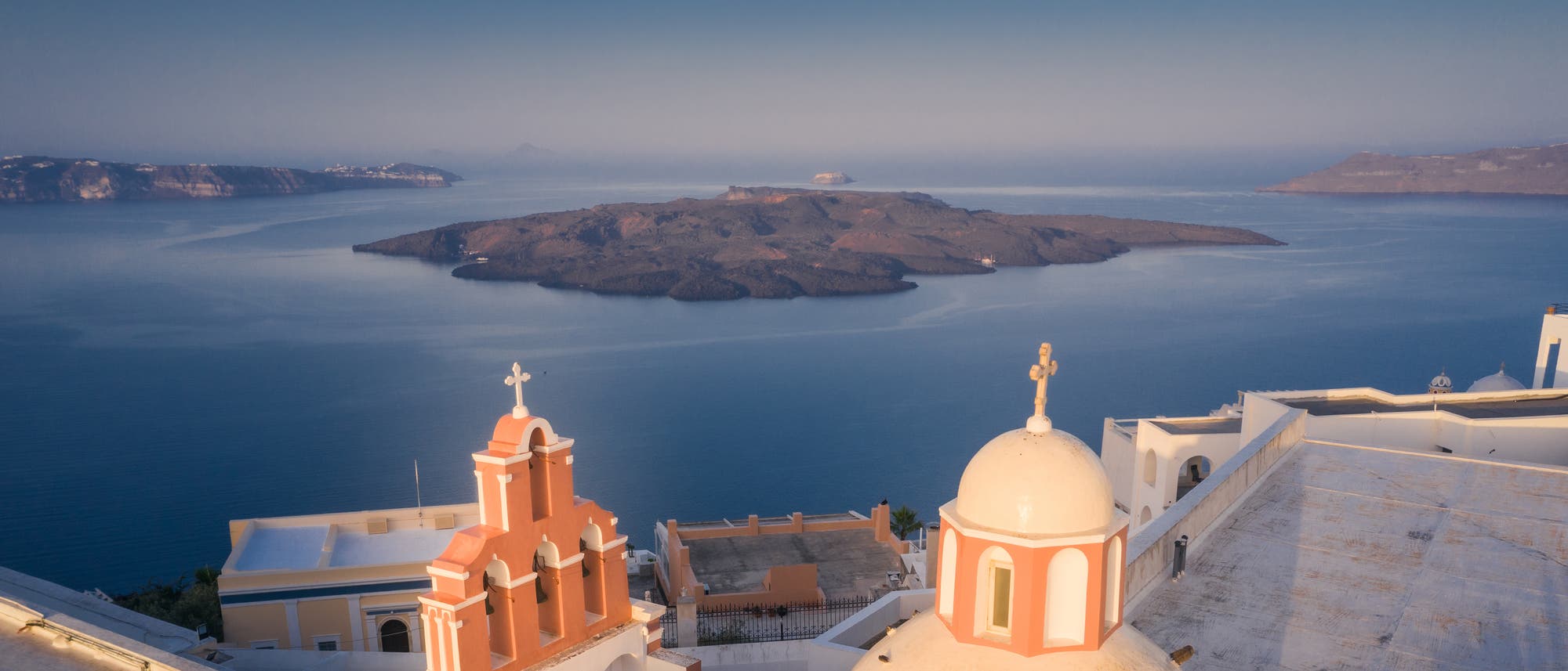 Blick auf die malerische Insel Santorini in Griechenland mit der charakteristischen Architektur der weißen Gebäude und Kuppeln im Vordergrund. Im Hintergrund ist die Vulkaninsel Nea Kameni im tiefblauen Meer zu sehen. Die Szene wird von einem klaren Himmel überragt, der die ruhige und idyllische Atmosphäre der Region unterstreicht.