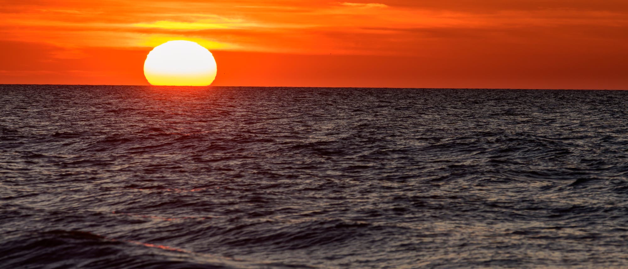 Sonnenuntergang über dem Meer mit einem leuchtend orangefarbenen Himmel. Die Sonne steht tief am Horizont und spiegelt sich im welligen Wasser. Keine Menschen oder Text im Bild. Sonnenuntergang über dem Meer mit einem leuchtend orangefarbenen Himmel. Die Sonne steht tief am Horizont und spiegelt sich im welligen Wasser. Keine Menschen oder Text im Bild.