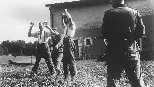 Schwarz-weiß-Aufnahme der Gefangennahme zweier Männer: im Vordergrund ist ein Soldat mit Gewehr zu sehen, das Gewehr ist auf die beiden mit erhobenen Händen dastehenden Männer gerichtet, ein weiterer Soldat nähert sich den gefangen genommenen von hinten.
