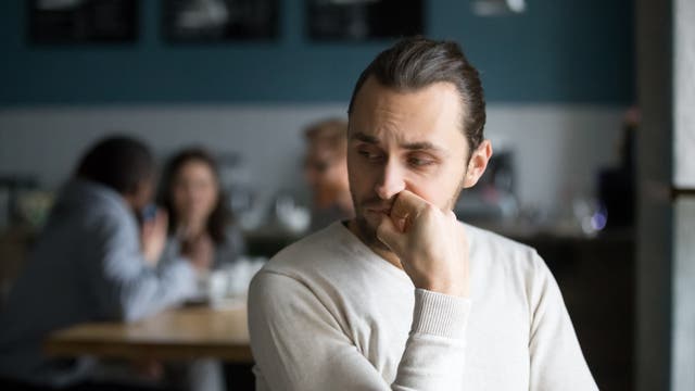 Ein etwas unglücklich dreinschauender Mann sitzt im Vordergrund. Er hat seinen Kopf auf eine Hand gestützt und leicht zur Seite gedreht, als würde er den Personen im Hintergrund lauschen, die dort im Gespräch an einem Tisch sitzen.