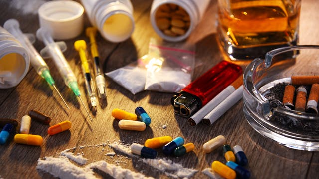 Eine Nahaufnahme eines Tisches mit verschiedenen Drogen und Utensilien. Zu sehen sind bunte Pillen, weiße Pulverlinien, Spritzen, Zigarettenstummel in einem Aschenbecher, ein Feuerzeug und ein Glas mit einer bernsteinfarbenen Flüssigkeit. Die Szene vermittelt ein Bild von Substanzmissbrauch und Sucht.
