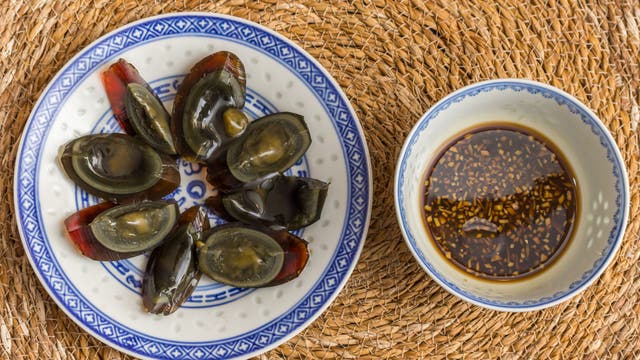 Ein Teller mit geschnittenen tausendjährigen Eiern auf einem blauen und weißen Porzellanteller, daneben eine Schale mit Sojasauce und Sesam. Die Speisen liegen auf einem geflochtenen Untersetzer. Traditionelle chinesische Delikatesse.