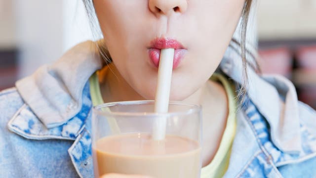 Eine Person in einer Jeansjacke trinkt einen Milchtee mit einem breiten Strohhalm aus einem Glas. Der Fokus liegt auf den Lippen und dem Strohhalm. Im Hintergrund sind verschwommene Details eines Cafés zu erkennen.