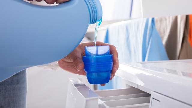 Eine Person gießt flüssiges Waschmittel aus einem großen blauen Behälter in die Kappe, um es in die Waschmaschine zu geben. Im Hintergrund sind bunte Kleidungsstücke zu sehen, die auf einer Leine hängen. Die Szene spielt in einem hellen Raum, der auf eine Waschküche hinweist.