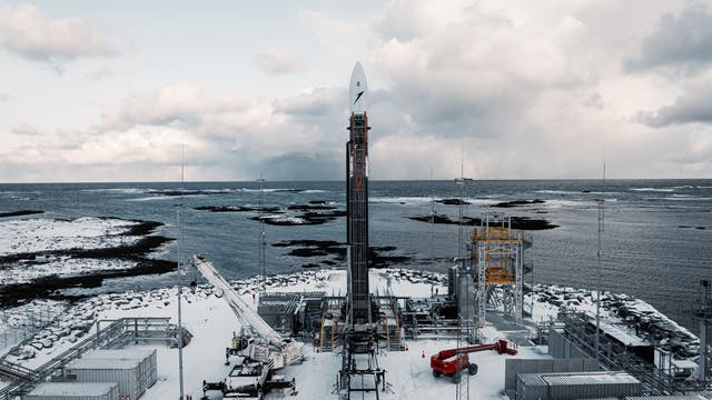 Eine Rakete steht auf einer verschneiten Startrampe an der Küste, umgeben von technischen Anlagen und Containern. Im Hintergrund ist das Meer mit kleinen Inseln zu sehen, darüber ein bewölkter Himmel. Die Szene vermittelt eine ruhige, aber technisch fortschrittliche Atmosphäre.