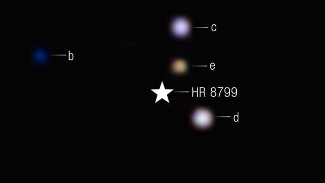 Ein Bild des Sternsystems HR 8799, das im Zentrum als weißer Stern dargestellt ist. Um den Stern herum sind vier Exoplaneten mit den Bezeichnungen b, c, d und e sichtbar. Planet b erscheint blau und ist links vom Stern positioniert. Die Planeten c, d und e sind rechts vom Stern und erscheinen in verschiedenen Farbtönen. Der Hintergrund ist schwarz.